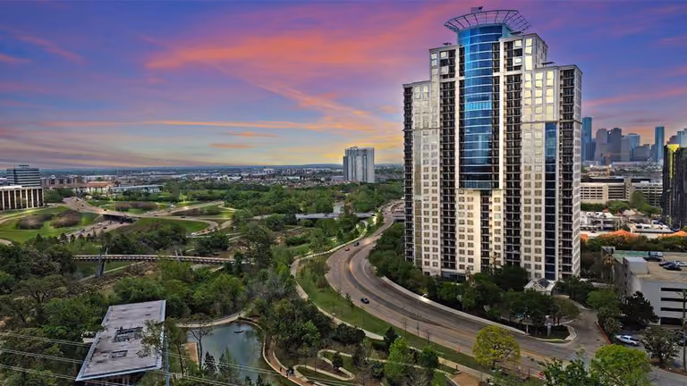 Aerial photograph at sunset of The Royalton at 3333 Allen Parkway: the tower stands at the right of the frame, a pale stone facade banded with dark balconies and a curved blue-glass bay rising through its centre, with Allen Parkway curving past its base. Parkland fills the left of the frame — mown lawns, walking trails, mature trees, a pond and a long pedestrian bridge over the bayou — and the downtown Houston skyline sits on the horizon under a pink and violet sky.