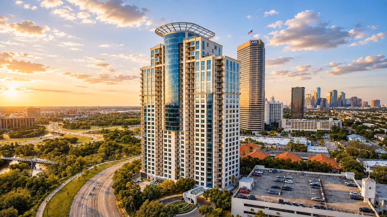 Aerial photograph of The Royalton at 3333 Allen Parkway in low golden light: the tower stands at the centre of the frame, a pale stone facade banded with balconies and a curved blue-glass bay rising through its centre to an open circular crown, with Allen Parkway curving past its base. A taller bronze-glass office tower rises just behind it, a parking structure and tile-roofed buildings fill the foreground to the right, and parkland — lawns, mature trees, a pond and a pedestrian bridge over the bayou — fills the left of the frame, with the downtown Houston skyline on the horizon under gold-lit clouds and the sun low at the far left.